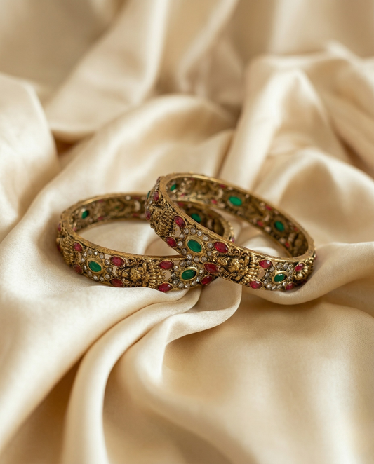Two ornate gold bangles with gemstones on a beige fabric background