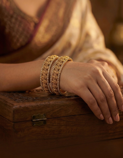 Close-up of a hand wearing gold bracelets with green stones on a wooden surface.