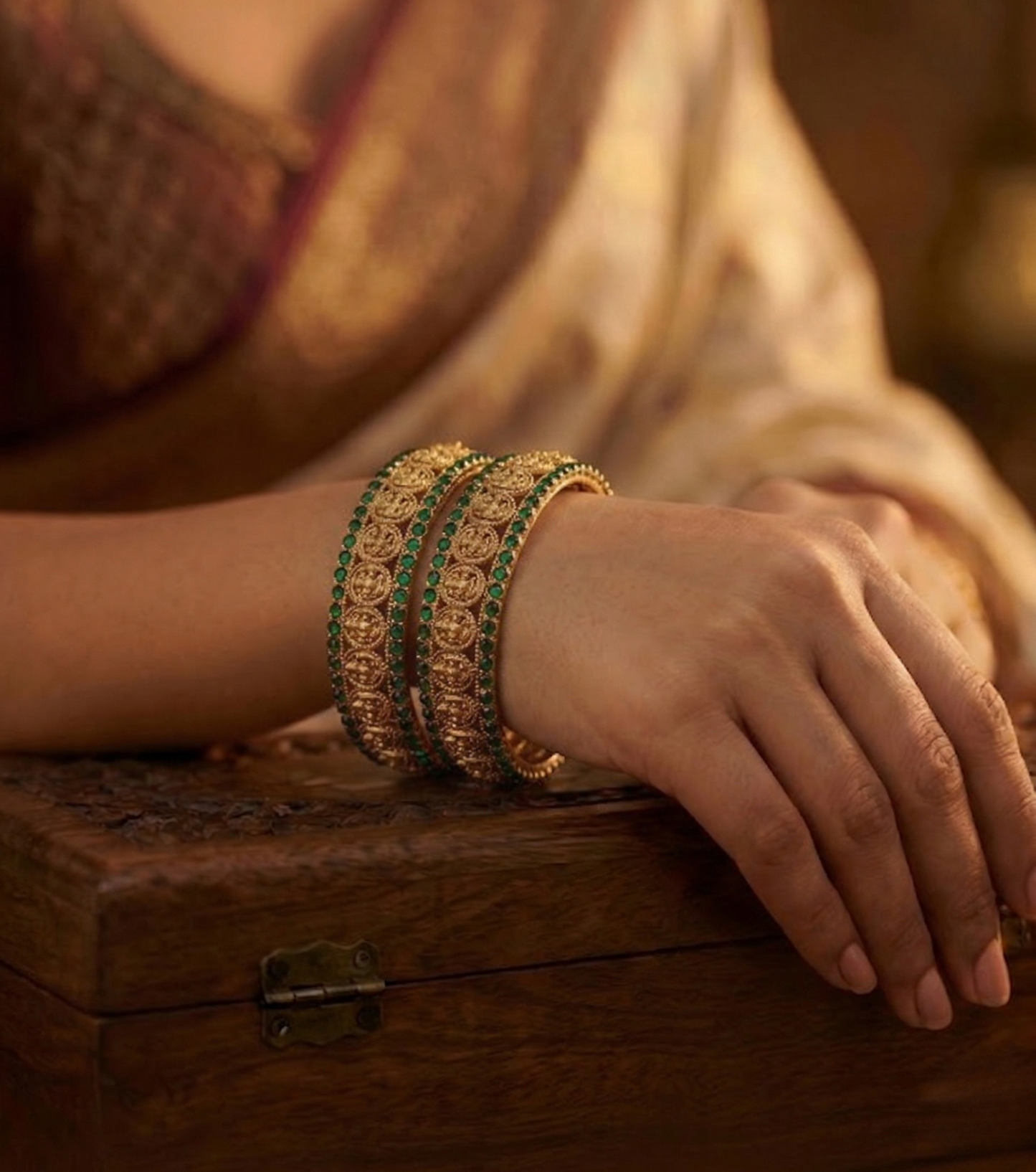 Close-up of a hand wearing gold bracelets with green stones on a wooden surface.