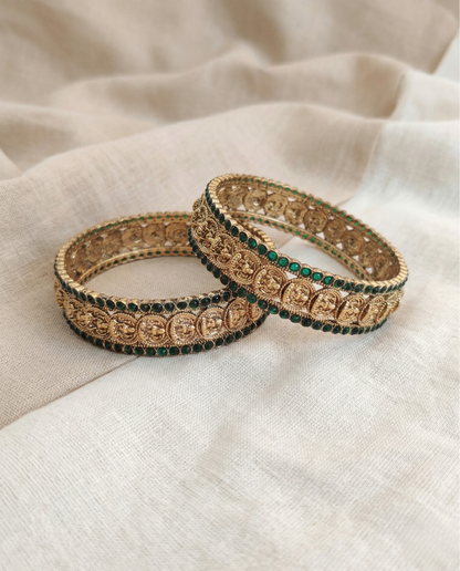 Two ornate gold and green bangles on a beige fabric background