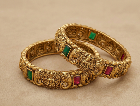 Pair of ornate gold bangles with green and red stones on a beige background