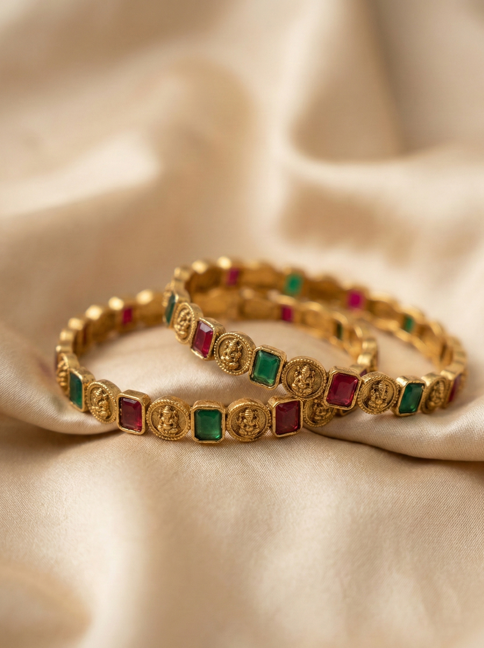 Gold bracelets with embedded red and green stones on a beige fabric background
