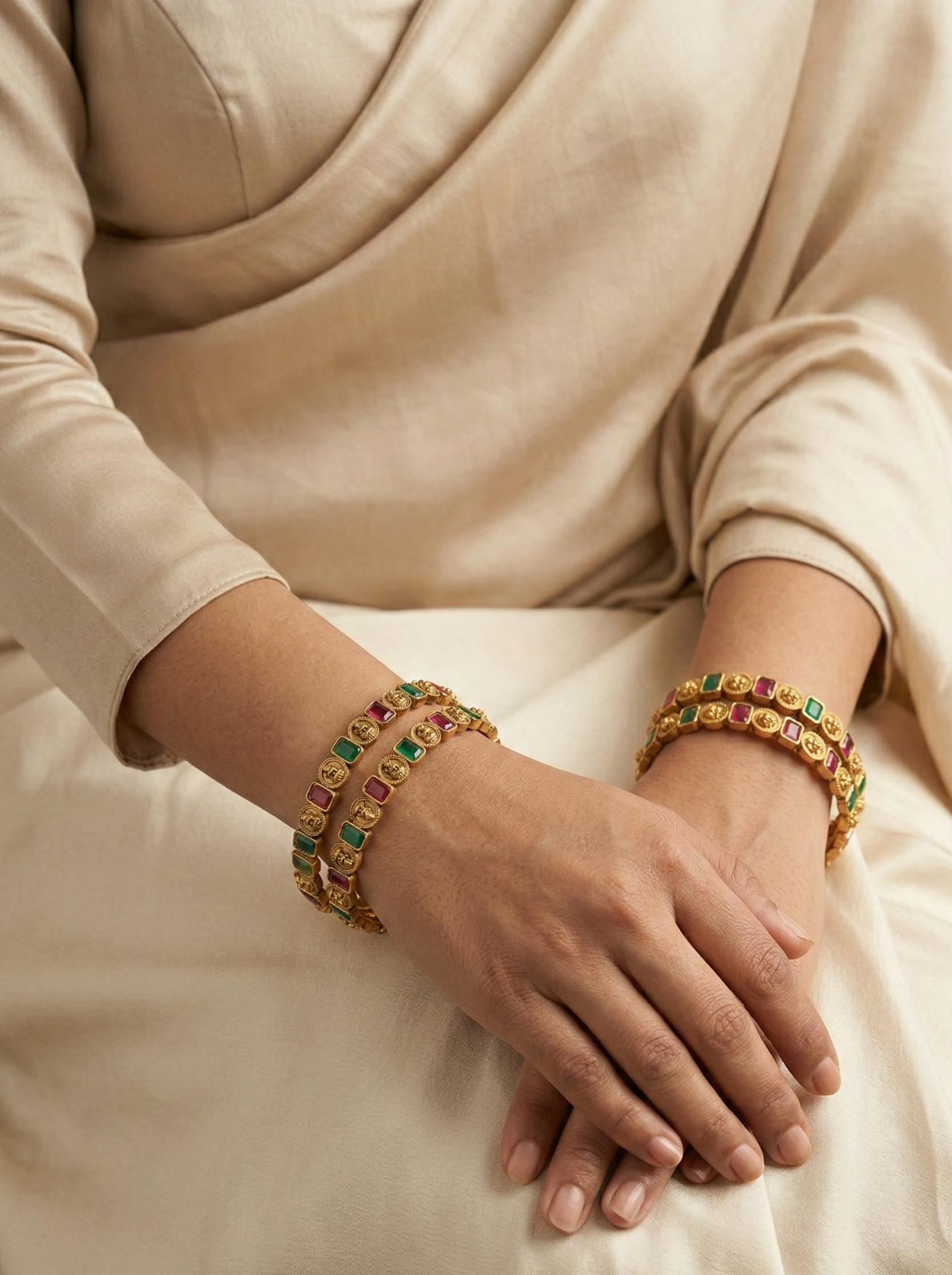 Close-up of hands wearing gold bracelets with gemstones on a light fabric background