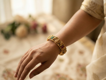 Hand wearing a gold bracelet with green and red stones, blurred floral background