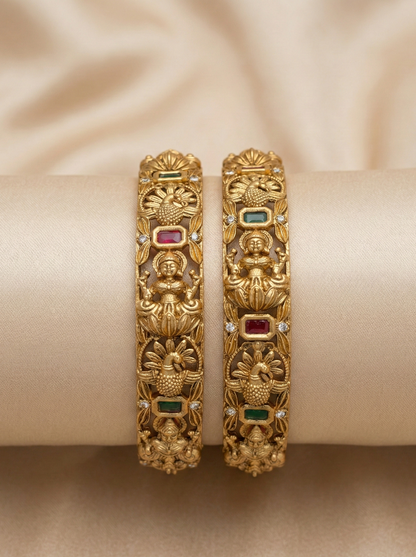 Pair of ornate gold bangles with gemstones on a beige fabric background