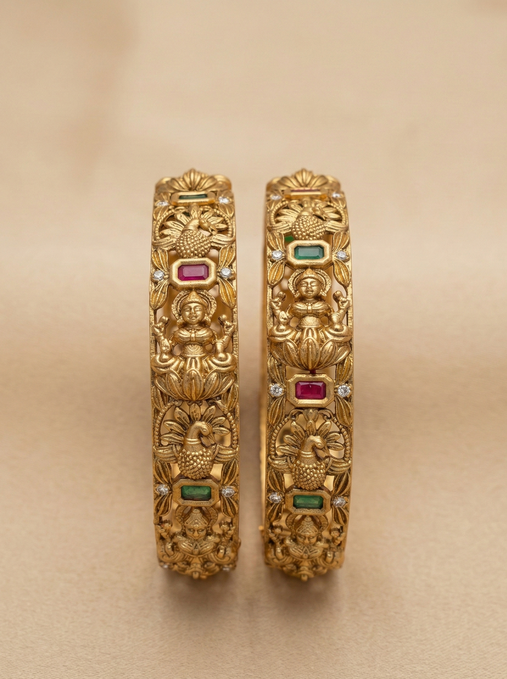 Pair of gold bangles with intricate designs and gemstones on a beige background