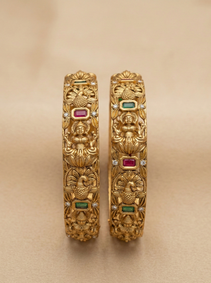 Pair of gold bangles with intricate designs and gemstones on a beige background
