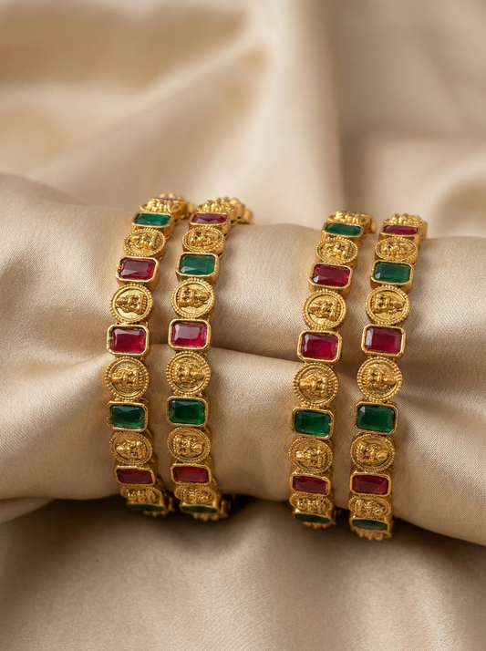 Gold bracelets with red and green gemstones on a beige fabric background