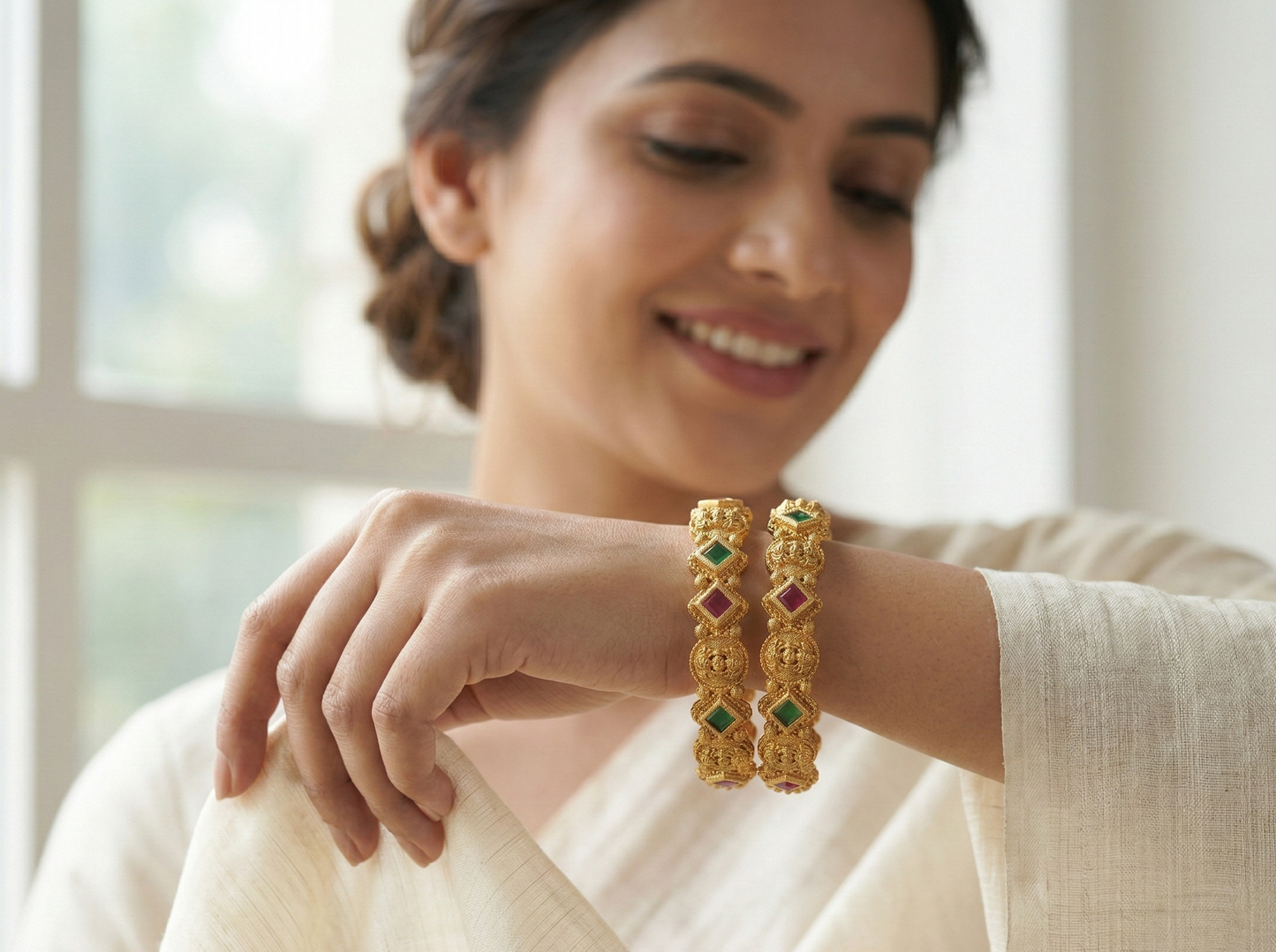 Woman wearing gold bangles with green stones, smiling indoors.
