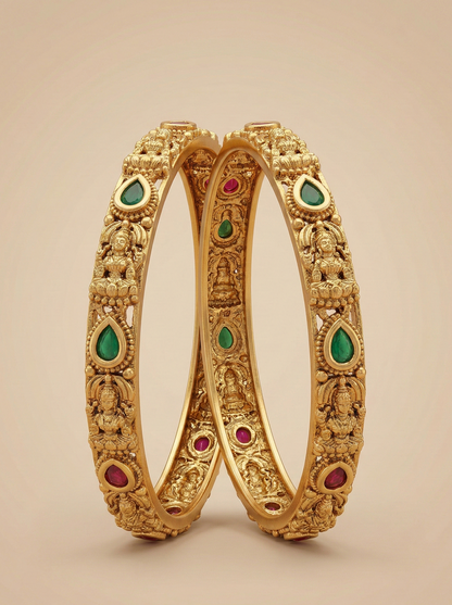 Pair of gold bangles with green and red gemstones on a beige background