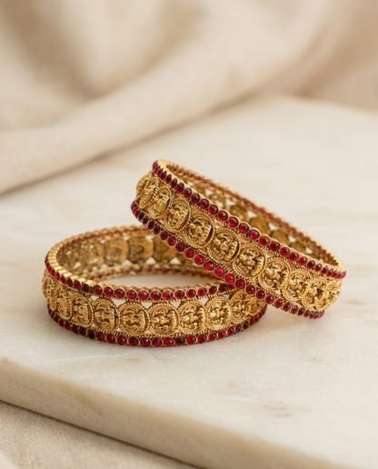 Two gold bangles with red stones on a marble surface