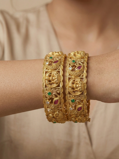 Gold bangles with gemstones on a person's wrist against a neutral background