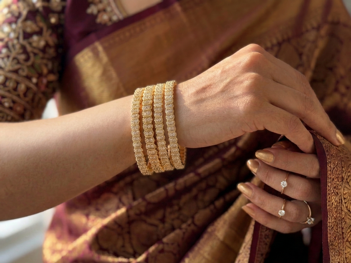 Close-up of a person wearing gold bangles and a ring, holding a brown saree.